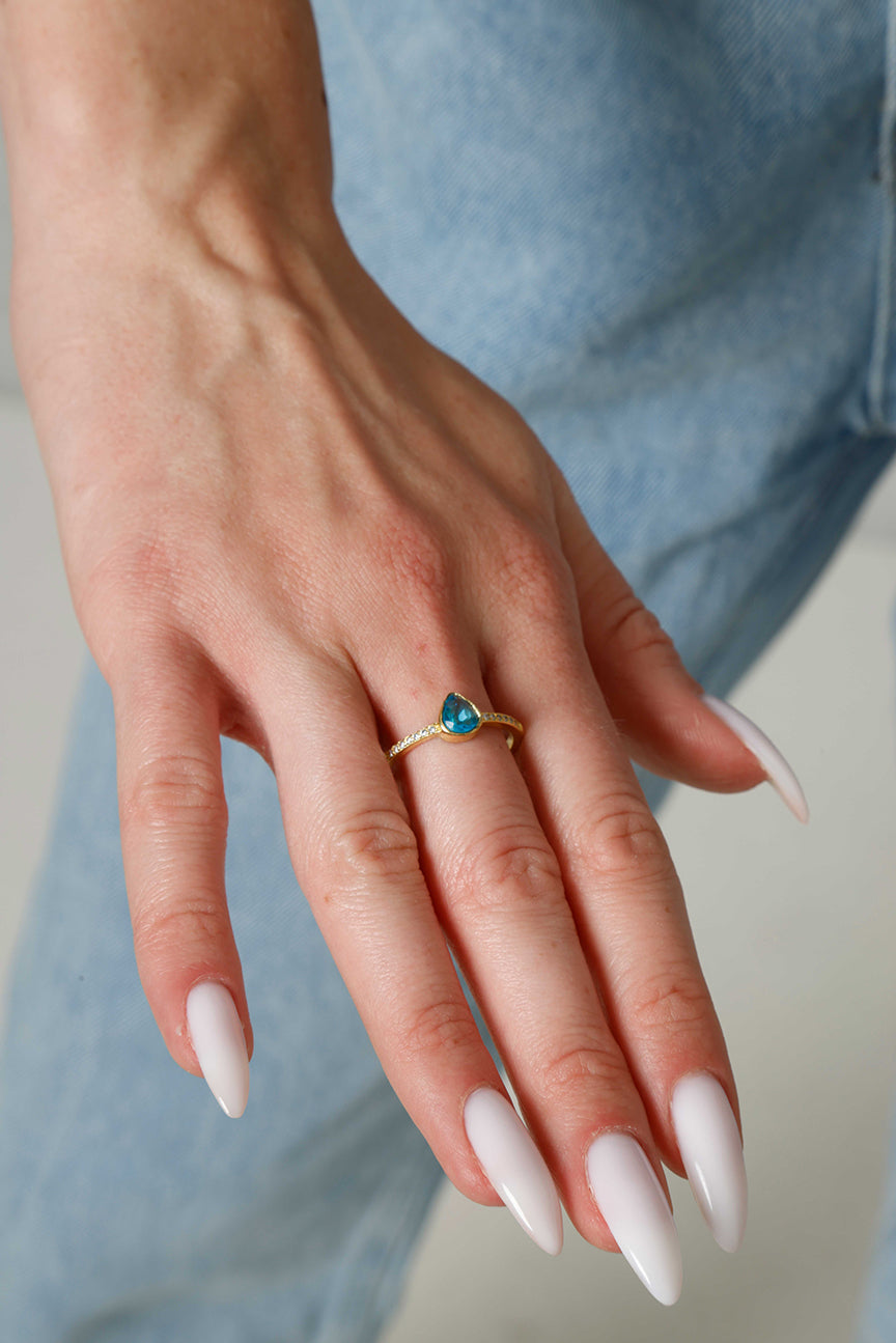 Hand wearing a gold ring with a blue gemstone, against a blurred denim background