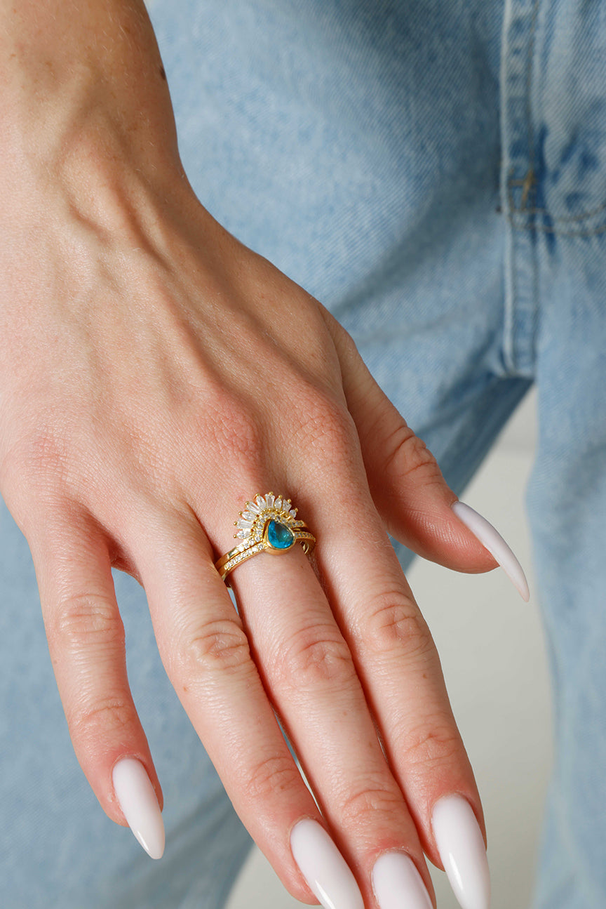 Hand wearing a gold ring with a blue gemstone against a light blue denim background