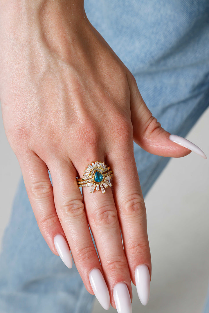 Hand wearing a stacking 
gold ring with a blue gemstone against a blurred background